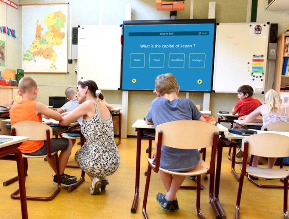 A teacher assisting students in learning with a Jeopardy game in a classroom, displaying a multiple-choice question on the projector screen